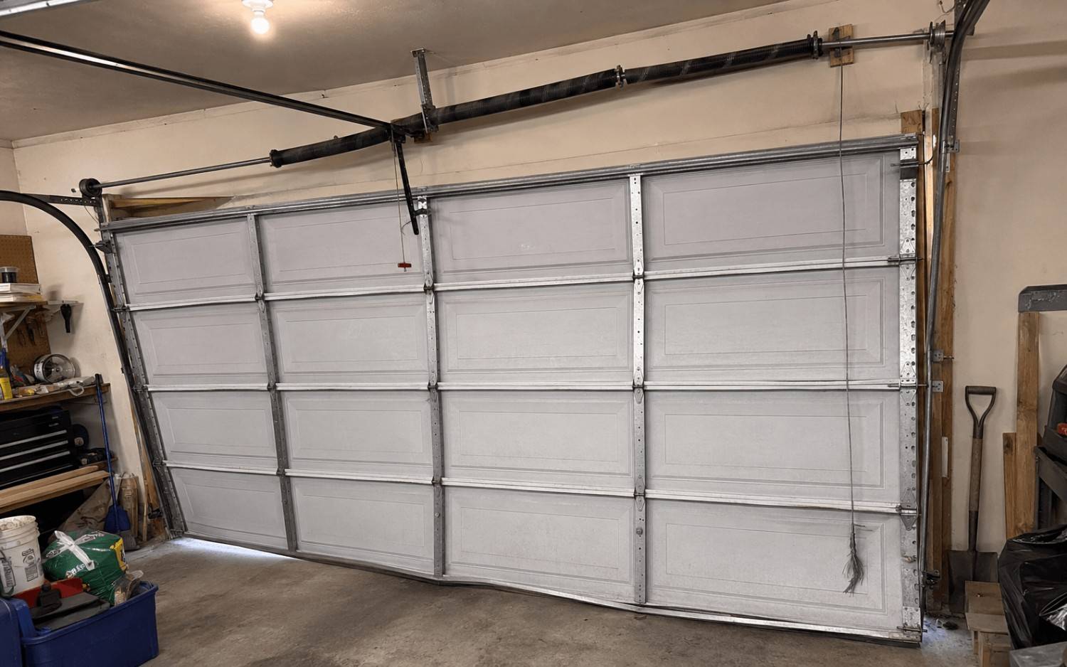 A garage door hanging crooked after a lift cable snapped, with the broken cable dangling down one side.